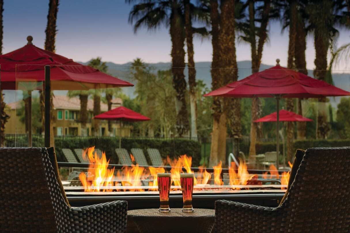 A fire feature with two glasses of a beverage, surrounded by wicker chairs and red umbrellas, with a view of palm trees and buildings in the background.