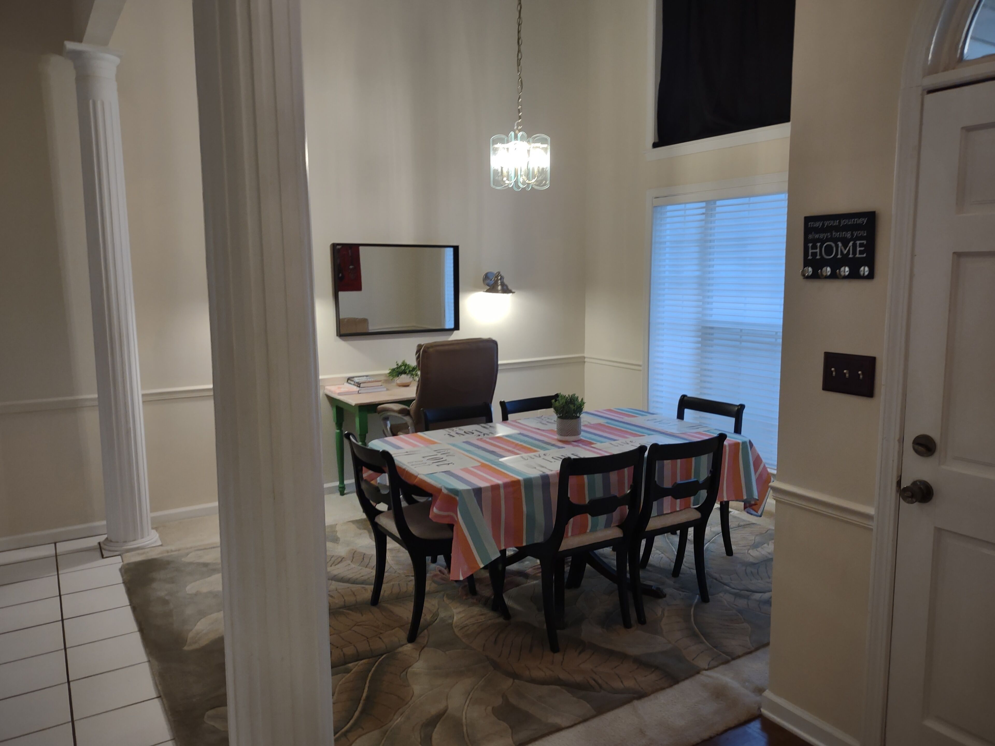 Un coin repas avec une table recouverte d’une nappe à rayures, entourée de chaises. Un lustre est suspendu au-dessus de la table, et un pot de fleurs est posé sur la table. Un miroir est accroché au mur au-dessus de la table, et une inscription sur le mur indique « MAISON ».