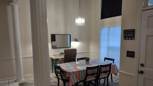 Een eethoek met een tafel bedekt met een geruit tafelkleed, omringd door stoelen. Daarboven hangt een kroonluchter, en op de tafel staat een potplant. Boven de tafel hangt een spiegel, en op de muur staat een bordje met de tekst 'HOME'.