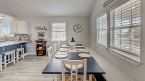 Un espace repas avec une longue table dressée pour six personnes, des chaises blanches et un lustre.