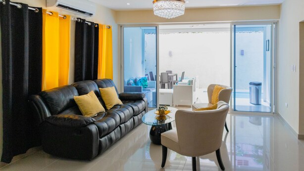 A modern living room with a black leather sofa, a glass coffee table, and a beige armchair. There is a chandelier hanging from the ceiling, and a sliding glass door leading to a balcony. - Mansión Versatil with a beautiful private pool 🏊‍♀️ 5 min from movies/groceries  (La Romana)