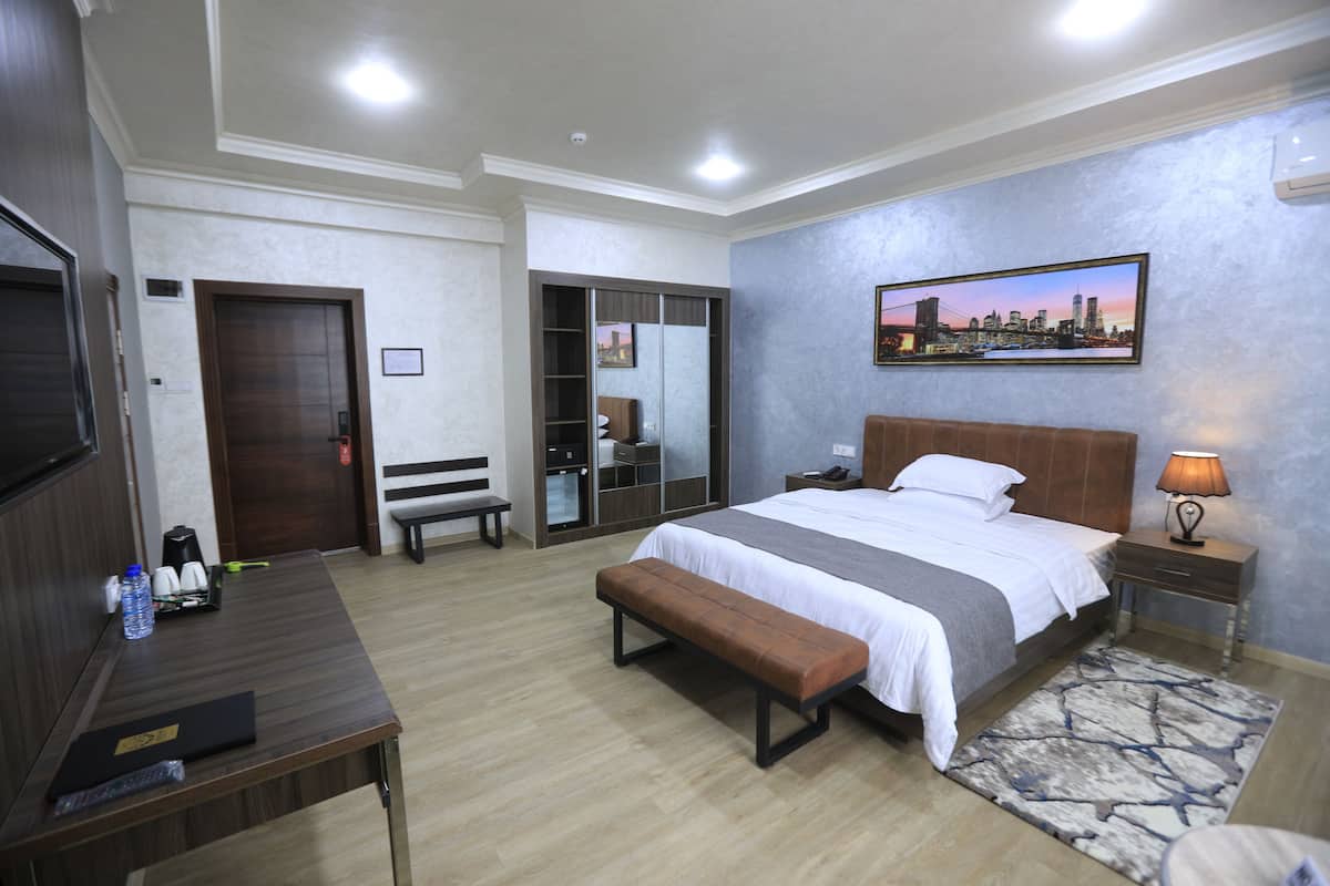 Une chambre d’hôtel avec un lit, une banquette, une table de chevet, une commode et un tableau représentant un paysage urbain accroché au mur.