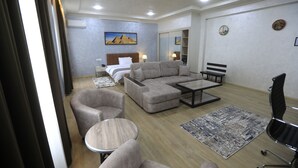 Et hotelværelse med en stor sofa, et rundt sofabord, et lille rundt bord, en sort kontorstol, en seng med hvid sengegavl, et natbord, en lampe, et maleri af pyramider, et spejl og et tæppe.