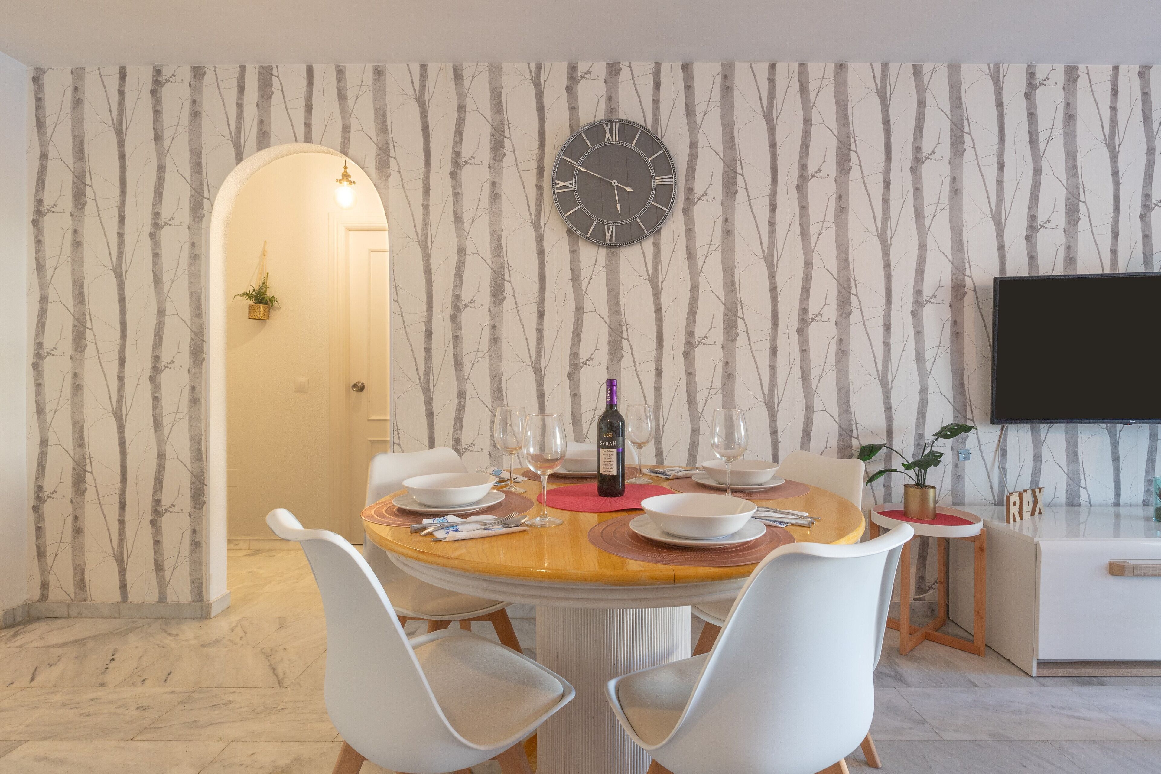 A dining area with a round table, white chairs, a wall-mounted clock, and a flat-screen TV.