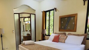 A well-lit hotel room with a large bed, a bedside table, a mirror, a window with curtains, and a wall-mounted air conditioner. - Enchanted Nest of Love! (Quepos)