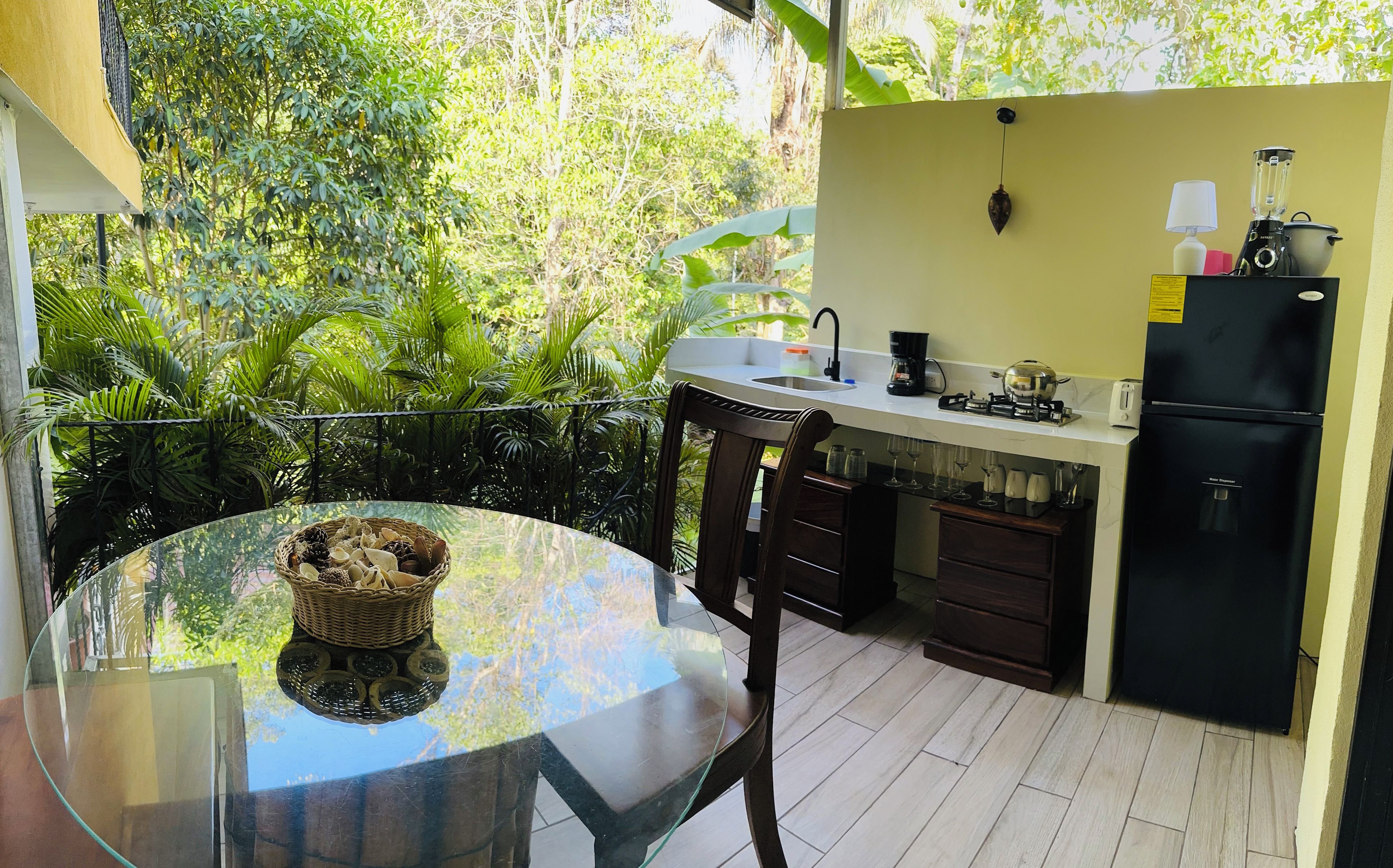 A small kitchen with a glass table, a wicker basket, and a view of lush greenery.