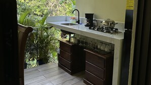 A compact kitchen with a sink, stove, and cabinets, overlooking a lush garden. - Enchanted Nest of Love! (Quepos)