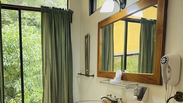 A bathroom with a wooden vanity, a glass bowl sink, a towel rack, a mirror, and a window with curtains. - Enchanted Nest of Love! (Quepos)