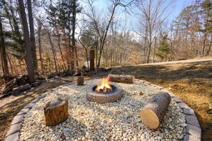 A circular fire pit surrounded by pebbles and logs, set in a forested area with trees and a dirt path.