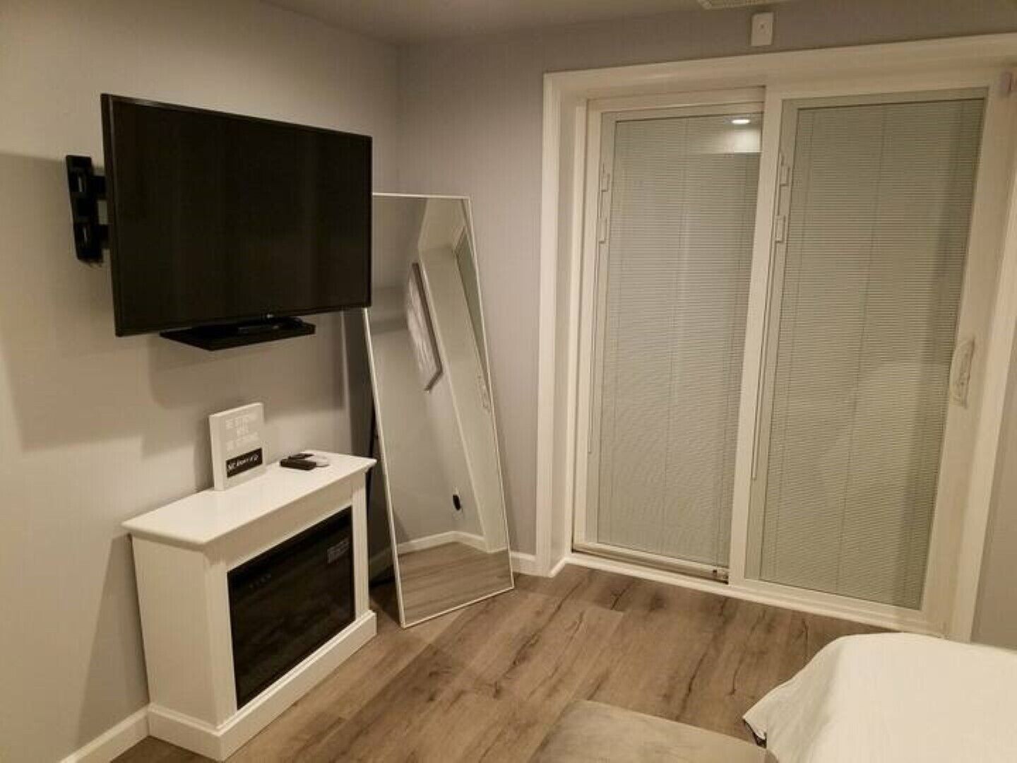 Une chambre avec un téléviseur fixé au mur, un placard avec une porte coulissante en verre et une table de chevet blanche.