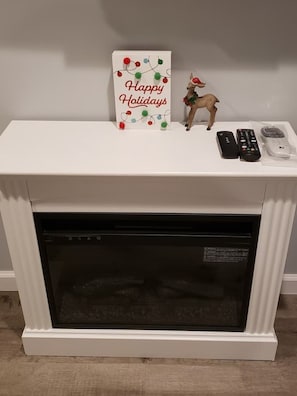 A fireplace with a white mantel, a 'Happy Holidays' sign, a small deer figurine, and two remote controls.