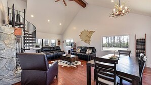 A spacious living room with a stone accent wall, a dining area with a wooden table and chairs, a fireplace, and a large window offering a view of the outdoors. - Secluded Lodge 8acres Hot Tub, Pingpong kayaks! (Sampson County)