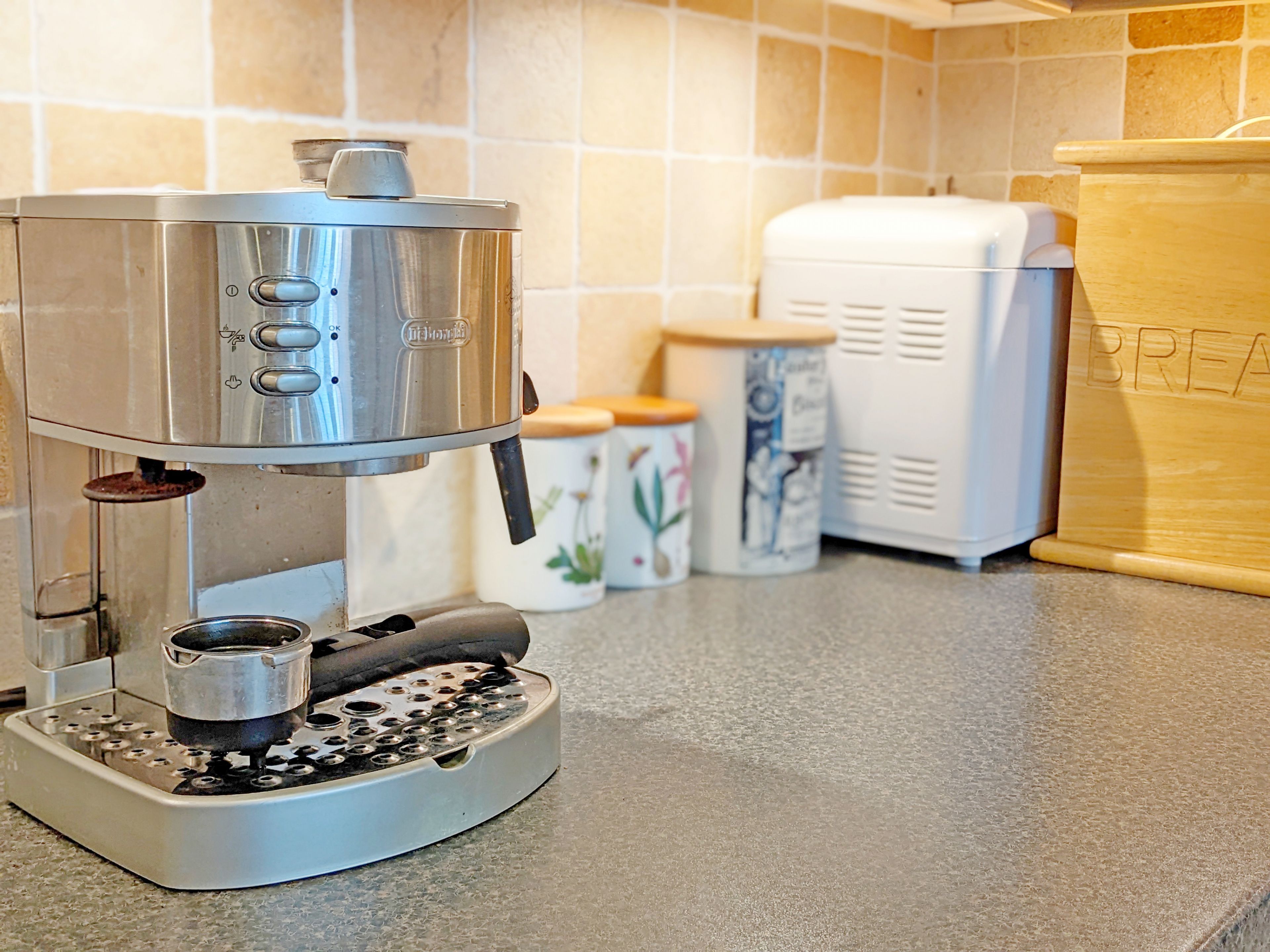 A coffee machine on a kitchen counter with a tiled backsplash and a bread box in the background.