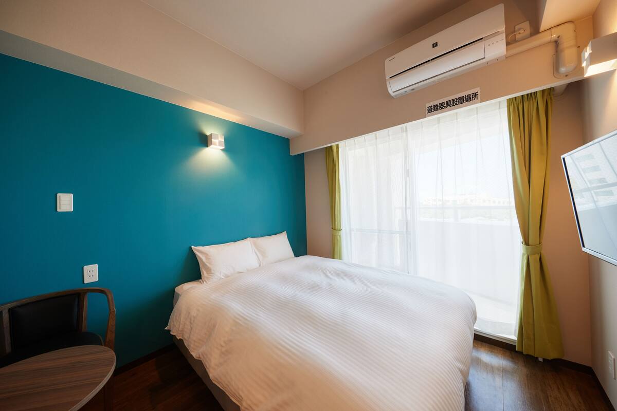 A hotel room with a bed, a desk, a chair, a window with curtains, and an air conditioning unit.