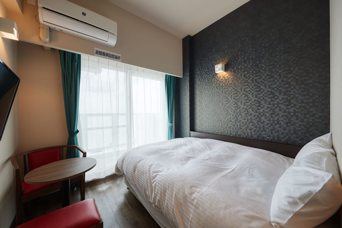 A hotel room with a bed, a small table, a red chair, and an air conditioning unit.