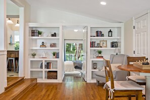 A cozy living room with a wooden floor, a dining table, and a bookshelf filled with books and decorative items.