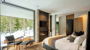 A modern bedroom with a large bed, a fireplace, a TV, and a view of a snowy landscape. - Kadenwood estate with heated pool & hot tub (Whistler)
