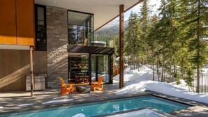 A modern outdoor pool area with a hot tub, seating, and a view of snow-covered trees. - Kadenwood estate with heated pool & hot tub (Whistler)