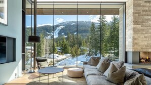 A modern living room with a large window, a fireplace, and a sofa. - Kadenwood estate with heated pool & hot tub (Whistler)