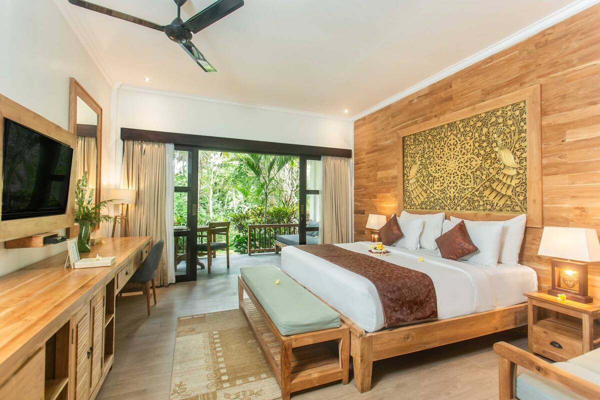 A bedroom with a wooden bed, a desk, a TV, and a balcony with a view of greenery.