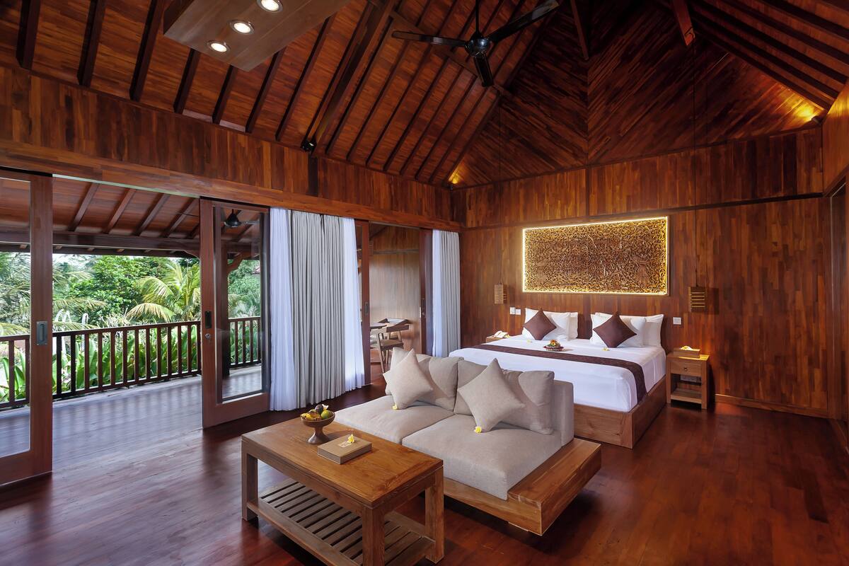 A spacious room with a large bed, a sofa, a wooden coffee table, and a balcony with a view of lush greenery.