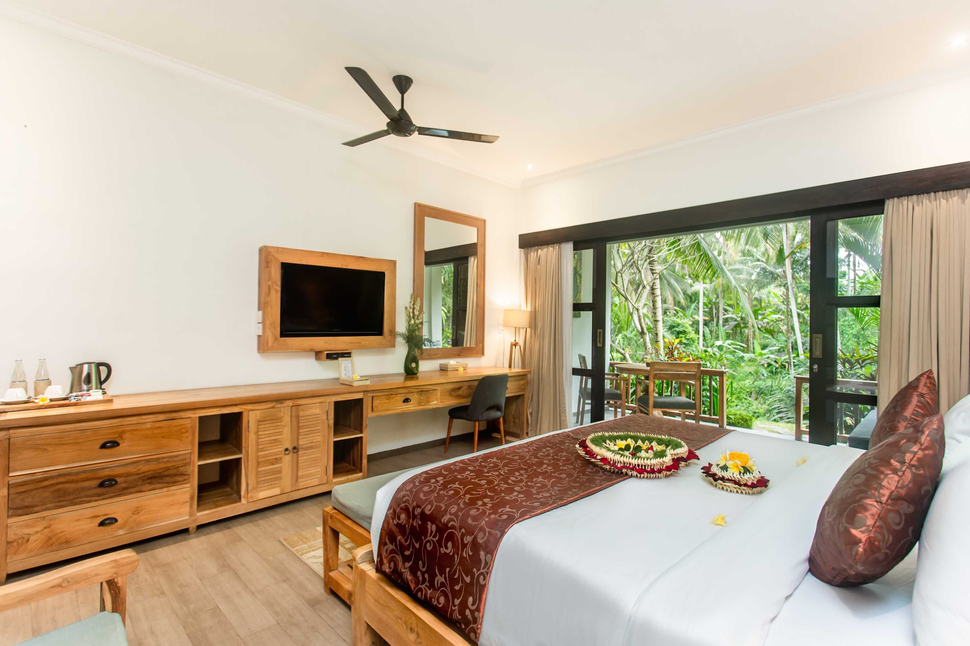 A hotel room with a bed, wooden desk, TV, and large windows with a view of greenery.