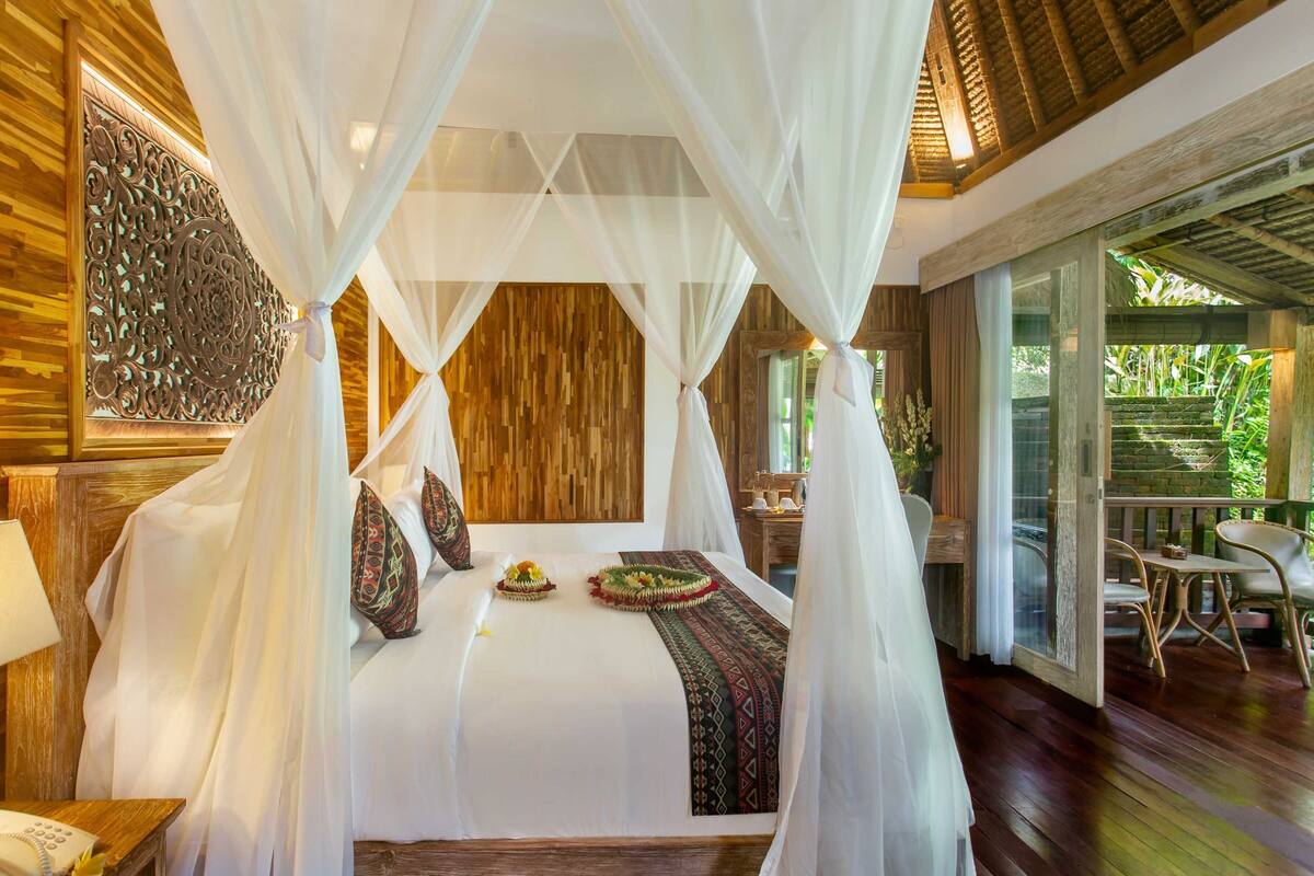 A room with a bed under a canopy, wooden walls, a small table with a lamp, and a view of greenery outside.