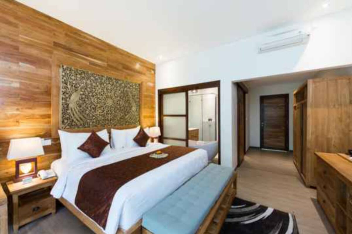 A modern hotel room with a wooden headboard, a large bed with white linens, a bedside table with a lamp, and a view of the hallway.