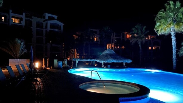 Ett område med en pool som är öppet på kvällen, komplett med bubbelpool, solstolar och palmer.