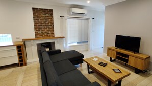 Een woonkamer met een hoekbank, een houten salontafel, een tv op een houten dressoir en een open haard met een bakstenen muur.