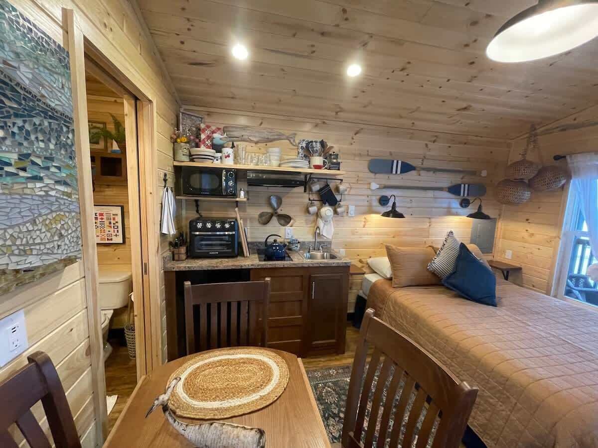 Interior acolhedor de cabana com mesa de jantar de madeira, cama com colcha acolchoada, área de cozinha com micro-ondas e diversos utensílios, e prateleira com louças e um objeto decorativo.