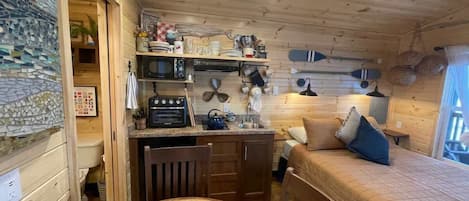 A cozy cabin interior with a wooden dining table, a bed with a quilted cover, a kitchen area with a microwave and various kitchen utensils, and a wall-mounted shelf with dishes and a decorative item.