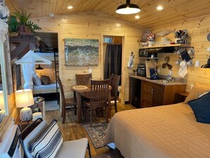 A cozy cabin-style room with a bed, a dining table with chairs, a kitchen area, and a living space with a couch and a lamp.