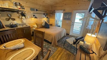 A cozy cabin interior with a wooden floor, a dining table with woven placemats, a bed with a blue pillow, and a small table with a lamp.