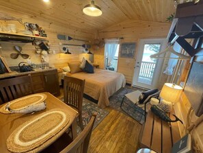 Interior acogedor de una cabaña con piso de madera, una mesa de comedor con individuales tejidos, una cama con almohada azul y una mesita de noche con lámpara.