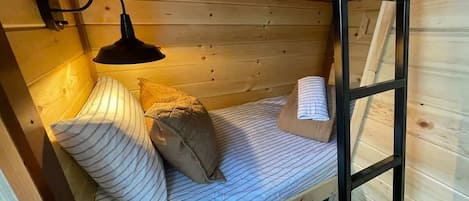 Una cama litera de madera con techo inclinado, una lámpara negra fijada a la pared y ropa de cama a rayas.