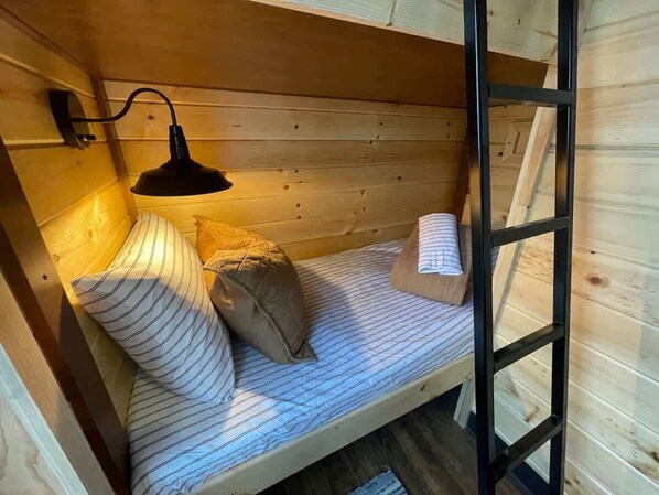 Una cama litera de madera con techo inclinado, una lámpara negra fijada a la pared y ropa de cama a rayas.
