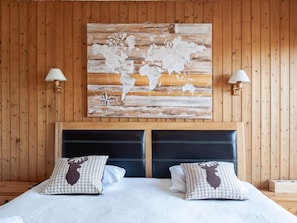 Quarto de hotel com parede revestida em madeira, uma grande ilustração de um mapa mundi, cabeceira preta e duas almofadas xadrez com desenho de alce.
