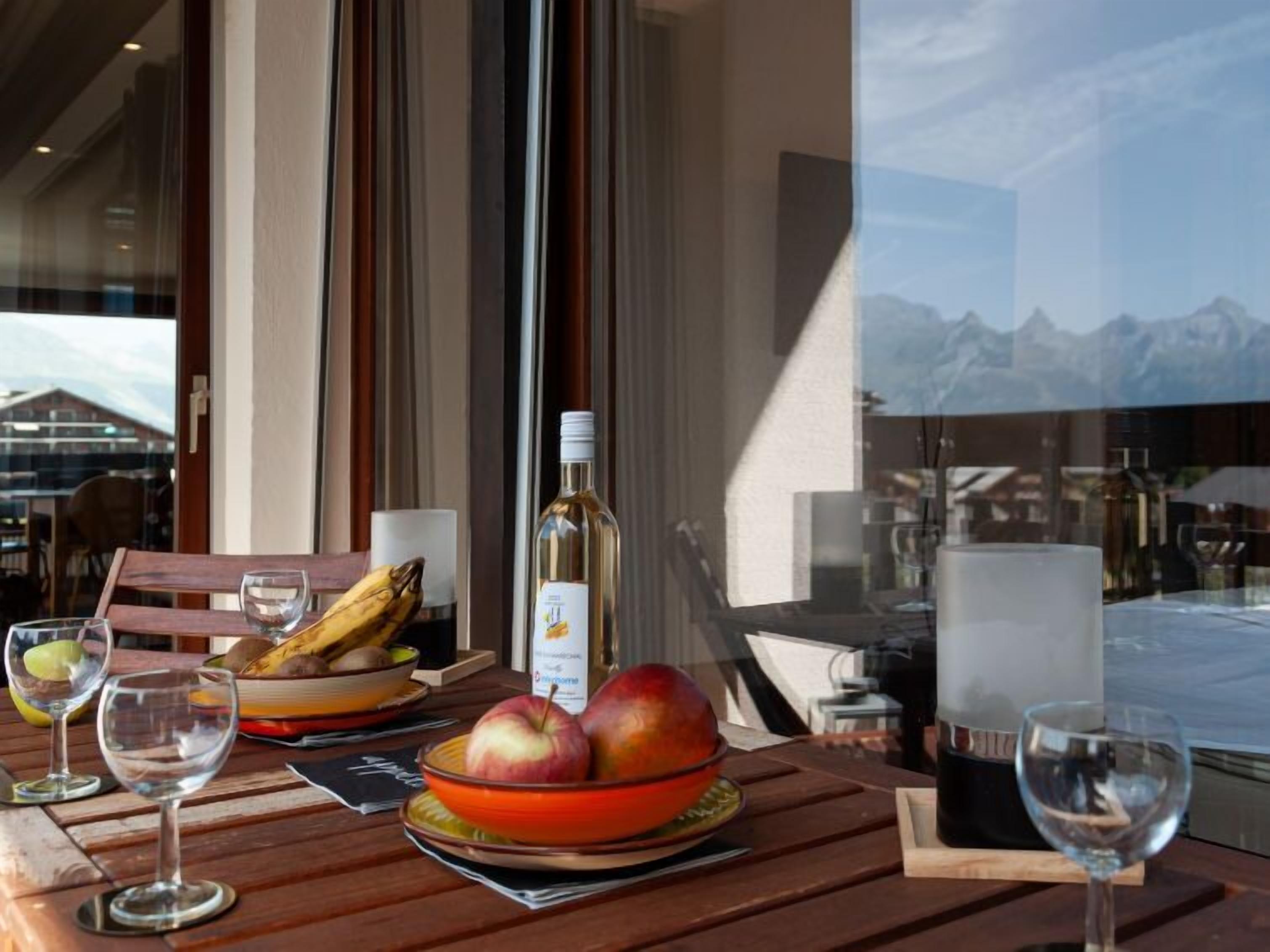 A table with a bottle of wine, a bowl of fruit, and glasses, set against a window with a mountain view.