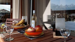 Une table avec une bouteille de vin, un saladier de fruits et des verres, le tout placé devant une fenêtre offrant une vue sur la montagne.