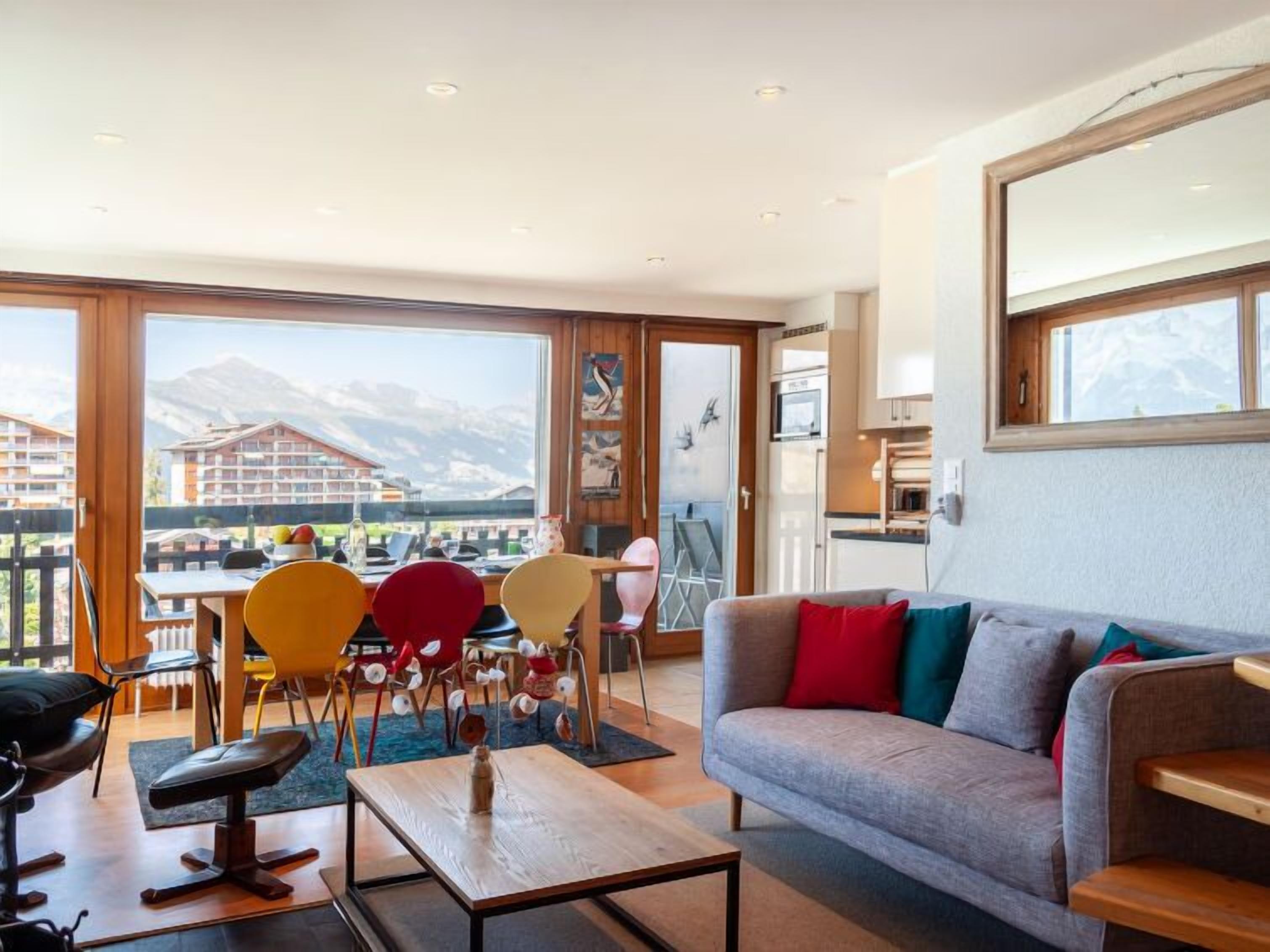 A modern living room with a sofa, coffee table, and chairs. A balcony with a view of buildings and mountains is visible through large windows.