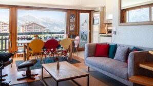 Un salon moderne avec un canapé, une table basse et des fauteuils. Un balcon avec vue sur les bâtiments et les montagnes est visible à travers de grandes fenêtres.