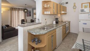 Cocina compacta con armarios de madera, horno empotrado y microondas. Cuenta con un comedor con mesa y sillas, y un sofá en la sala de estar contigua.