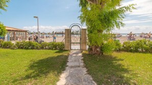 A pathway leading to a beach area with a gate, a tree, and a building with a roof. - Carabela Front Beach & Pool by Interhome (Port d'Alcúdia)