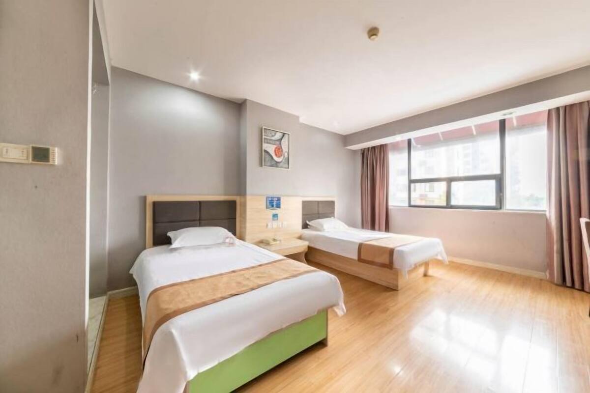 Ein Hotelzimmer mit zwei Betten, einem Nachttisch, einem Wandthermostat und einem Fenster mit Vorhängen.