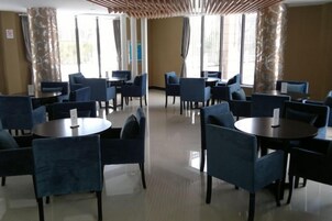 Un hall de hotel con mesas y sillas redondas, ventanas amplias y suelo de baldosas.