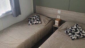 A twin room with two beds, a window with curtains, a shelf with a 'HAPPY' sign, and a bedside table with a lamp. - Zenitude AZ75 (Frejus)