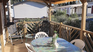 A covered outdoor patio with a wooden deck, white plastic chairs, a table with a pineapple-patterned tablecloth, and a small decorative plant. - Zenitude AZ75 (Frejus)