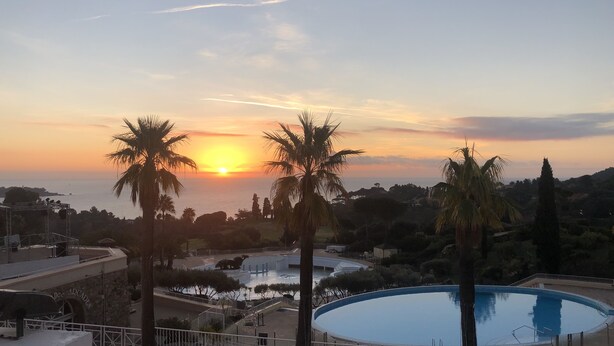 A sunset view from a poolside area with palm trees, a pool, and a distant body of water. - Cap Esterel Agay - large garden - exceptional sea and pool views (Saint-Raphaël)
