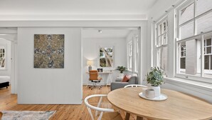 A modern living room with a wooden dining table, white chairs, and a large abstract painting on the wall. - Mulwarree Avenue Apartments by Urban Rest (Randwick)